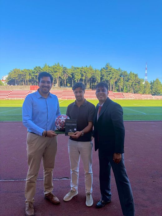 Jai Agarwal and two colleagues holding a signed football and framed certificate on the stadium track