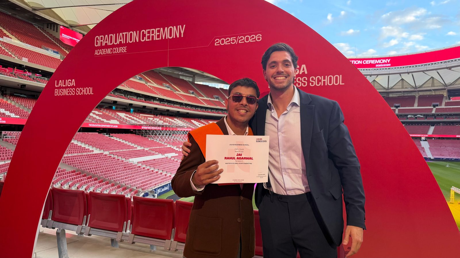 Jai Agarwal in an orange graduation stole holding his LaLiga Business School diploma, beside Alejandro Sierra Capitán, under the 2025/2026 graduation ceremony arch